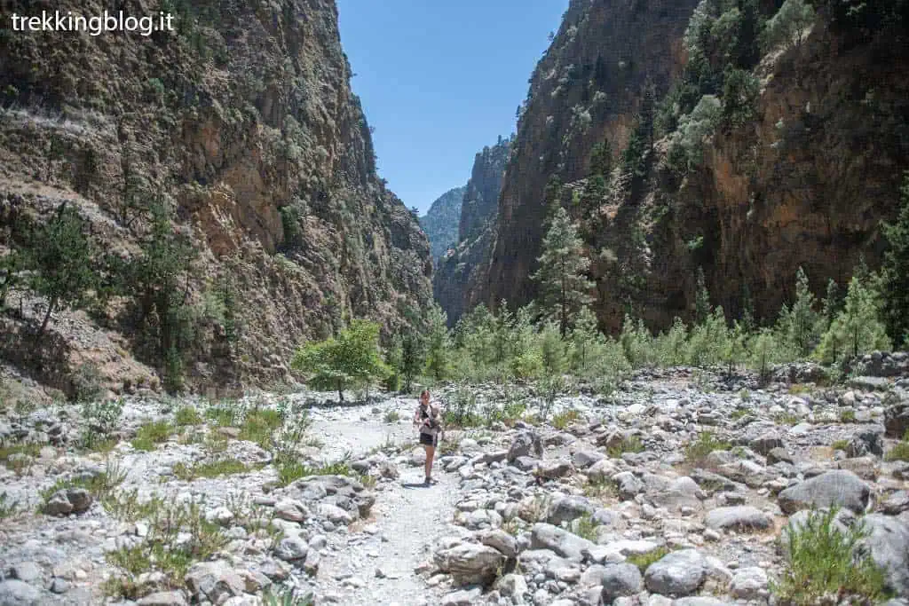 Una donna che cammina su un sentiero roccioso con la Gola di Samariá sullo sfondo